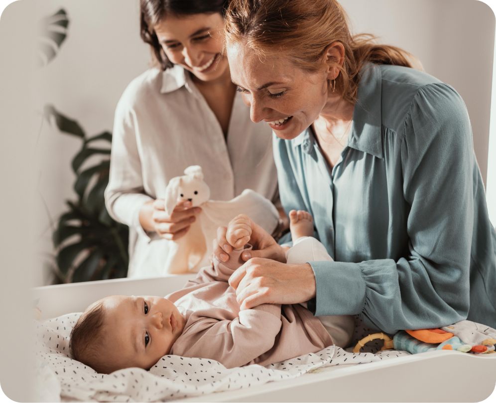 Baby auf Wickeltisch, dahinter stehen zwei lächelnde Frauen