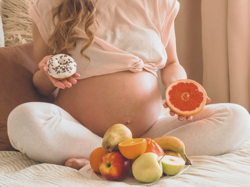 Schwangere Frau sitzt auf dem bett mit Obst in der einen Hand und einem Donut in der anderen Hand