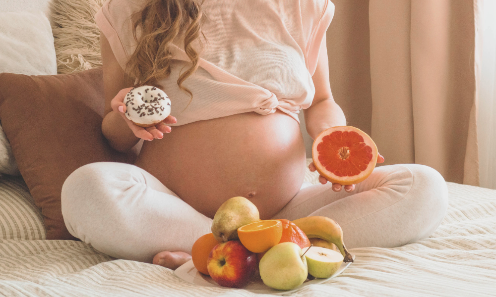 Schwangere Frau sitzt auf dem bett mit Obst in der einen Hand und einem Donut in der anderen Hand
